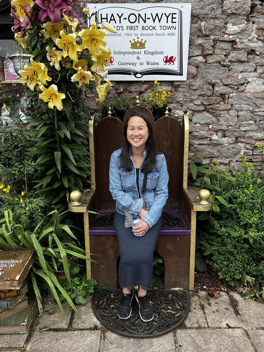 Smiling in a decorative throne-style chair beneath a sign reading Hay-on-Wye, World&rsquo;s First Book Town, Gateway to Wales